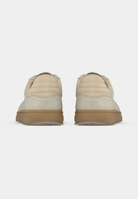 Beige suede sneakers with a textured tan rubber sole, featuring a padded heel and minimalistic design. Model displays a rear view.