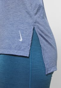 Camicia azzurra a maniche corte con fessure ai lati, caratterizzata da un piccolo logo Nike bianco. Indossata sopra leggings sportivi color teal.