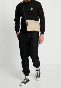 Beżowa torebka na ramię z zapięciem na zamek i regulowanym paskiem, wykonana z trwałego materiału. Posiada przedni logo oraz teksturowaną powierzchnię.