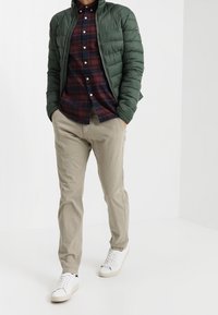 Grüne Steppjacke, marineblau-rotes kariertes Hemd, beige Hose und weiße Turnschuhe, mit glatter Textur und figurbetontem Design.