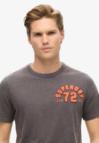 Camiseta de algodón gris con cuello redondo, que presenta el texto bordado en naranja "SUPERDRY 72 ATH" en el área del pecho izquierdo.