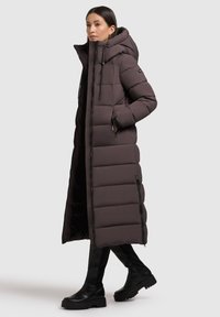 Dolga rjava puhasta jakna z veliko kapuco, prešita zasnova, zadrga spredaj in stranskimi žepi. Kombinirana s črnimi škornji do kolen.