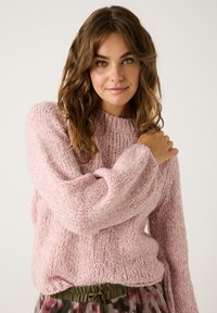 Rosa gestrickter Pullover mit strukturierter Oberfläche, rundem Halsausschnitt und langen Ärmeln. Kombiniert mit olivgrünen gemusterten Hosen.