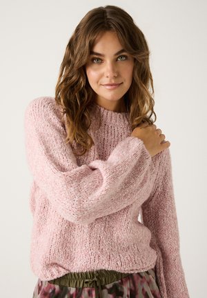 Rosa gestrickter Pullover mit strukturierter Oberfläche, rundem Halsausschnitt und langen Ärmeln. Kombiniert mit olivgrünen gemusterten Hosen.