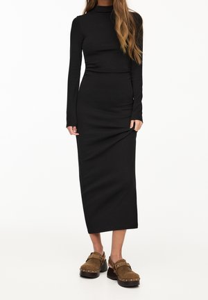 Femme portant une robe noire à manches longues et col montant, longueur midi, associée à des sabots marron cloutés, sur un fond uni.