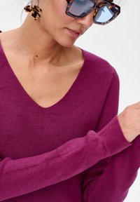 Dopasowany, żebrowany sweter w głębokim magencie z dekoltem w kształcie V. Miękka tekstura i długie rękawy. Przesadzone okulary przeciwsłoneczne w kolorze żółwia jako dodatek.