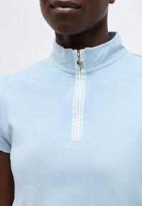 Κοντινό πλάνο ενός ατόμου που φοράει ανοιχτό μπλε κοντομάνικο με φερμουάρ με λευκή επένδυση, μισάνοιχτο στο γιακά.