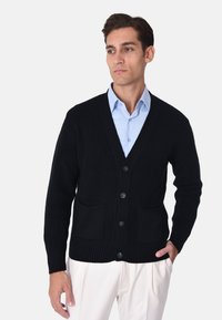Cardigan nero lavorato a maglia con scollatura a V, cinque bottoni, polsini a costine e due tasche frontali. Indossato sopra una camicia azzurro chiaro.