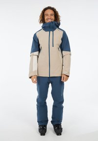 Beige en marineblauwe skijas met een hoge kraag, twee ritszakken en lange mouwen, gecombineerd met bijpassende marineblauwe skibroek en zwarte skiboots.