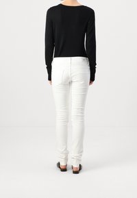 Personne portant un haut noir à manches longues et un jean blanc ajusté, debout pieds nus sur un fond blanc uni, tourné le dos au spectateur.