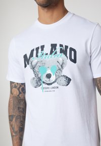White cotton t-shirt featuring a graphic of a grey teddy bear with blue paint accents. Text reads "MILANO Italia" and "Closure London."