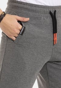 Graue Sweatpants mit einem Kordelzugbund, Seitentaschen und einem Textillabel. Mit orangefarbenen Akzenten an den Kordelzügen. Weiches Material.
