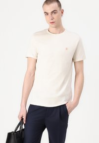 Tricou din bumbac bej, cu mâneci scurte, guler rotund și un mic logo portocaliu "II" pe piept, asortat cu pantaloni bleumarin și o geantă neagră.