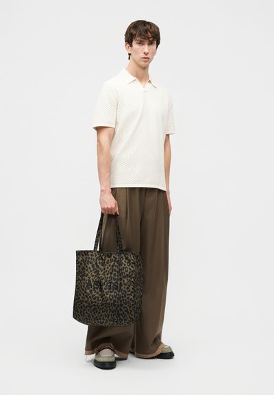 Jeune homme portant une chemise blanche texturée à manches courtes et un pantalon large marron, tenant un sac fourre-tout imprimé léopard avec un texte noir.