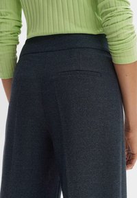 Pantalon bleu marine foncé en tissu texturé avec une coupe ajustée, doté d'une poche arrière unique. Porté avec un haut à côtes vert clair.