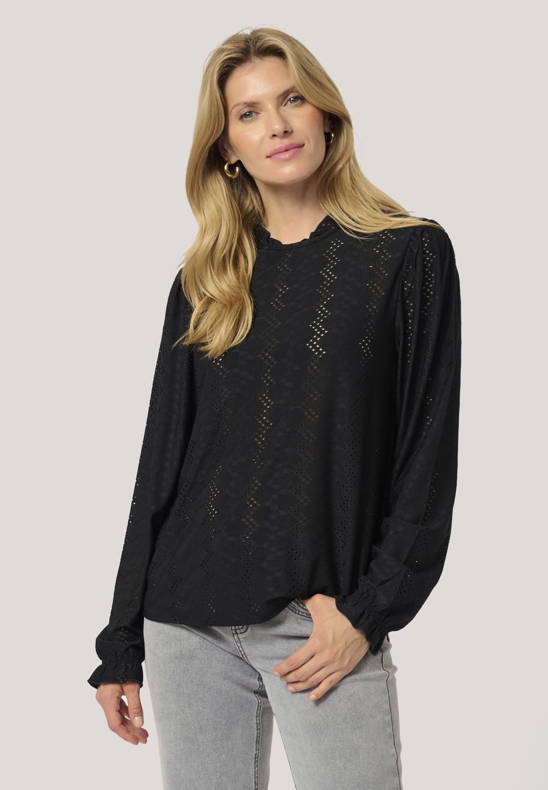 Blusa nera a maniche lunghe con dettagli di occhiello, scollatura volant e polsini elasticizzati. Tessuto morbido con una vestibilità rilassata. Abbinata a jeans grigio chiaro.