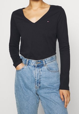 Persoon die een zwart langärmelig V-hals shirt draagt met een klein logo op de borst, ingestopt in een lichtblauwe high-waisted spijkerbroek.