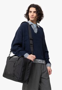 Sac messager noir de forme rectangulaire, avec une poche avant et une bandoulière réglable, tenu par une personne portant un pull en laine cardi bleu marine.