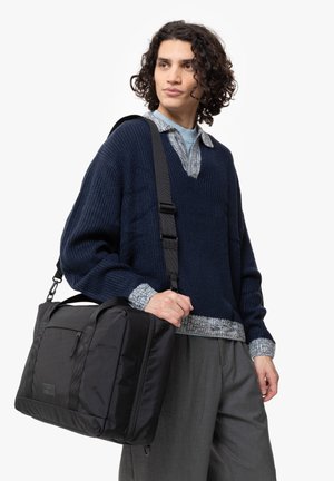 Black messenger bag with a rectangular shape, featuring a front pocket and adjustable strap, held by a person wearing a navy knit sweater.