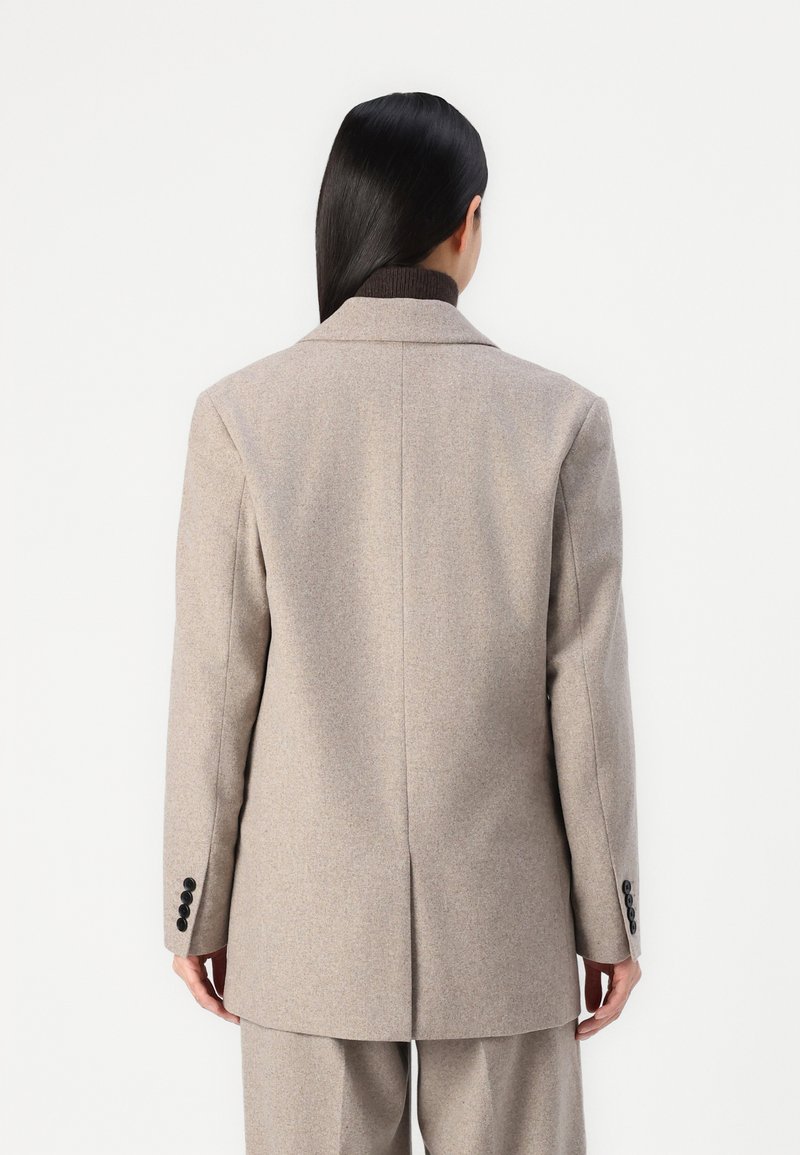 Donna con lunghi capelli scuri indossa un blazer beige su misura e pantaloni abbinati, in piedi di spalle contro uno sfondo bianco semplice.