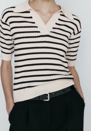 Persoon draagt een geribd, kort mouw crème topje met zwarte horizontale strepen, ingestopt in een zwarte broek met een zwarte riem en zilveren gesp.