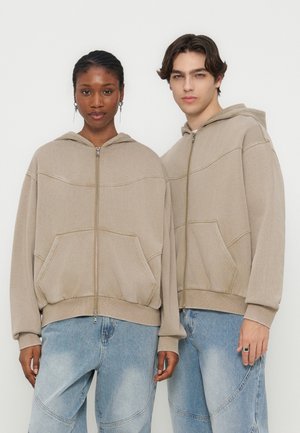 Dwie osoby ubrane w beżowe bluzy zapinane na zamek i jasnoniebieskie dżinsy, stojące obok siebie na jednolitym białym tle.