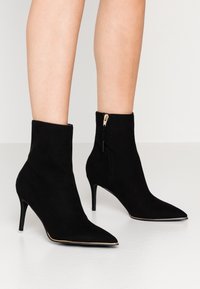 Jambes portant des bottines en daim noir à bout pointu avec des fermetures éclair latérales et des talons hauts fins sur un sol blanc.