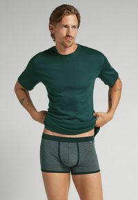 Man med mörkgrön t-shirt och grå boxershorts, som justerar sin skjorta, står mot en enkel ljus bakgrund.