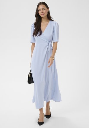 Femme aux cheveux longs et foncés portant une robe portefeuille bleu clair, des talons noirs, un sac à main noir et un bracelet en or.