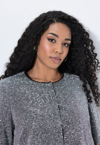 Vrouw met lang, krullend haar, draagt ringoorbellen en een gestructureerde zilvergrijze jas voor een effen lichte achtergrond.