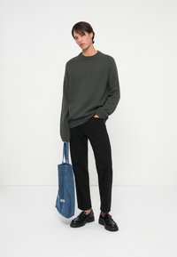 Ciemnozielony sweter z dzianiny, czarne spodnie i czarne buty. Trzyma niebieską torbę dżinsową z uchwytem, z białym tekstem po bokach.