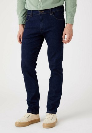 Man draagt donkerblauwe slim-fit jeans, lichtgroene blouse met knopen en gebroken witte sneakers met gumzolen, staand tegen een effen achtergrond.