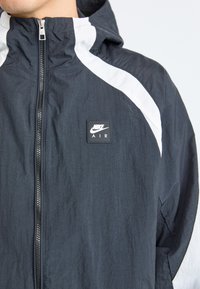 Nike Air -takki, jossa on vetoketjullinen edestä suljettava osa, laivastonsinistä kangasta, valkoiset verkkokoristeet olkapäillä ja musta logoplässi rinnassa.