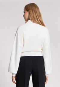 Biały sweter z dzianiny o prążkowanej strukturze, z wysokim kołnierzem i bufiastymi rękawami, w zestawieniu z czarnymi spodniami, widok z tyłu.