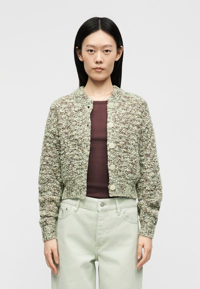 Femme portant un cardigan boutonné texturé vert-gris sur un haut bordeaux, assorti à un pantalon clair taille haute, se tenant devant un fond uni.