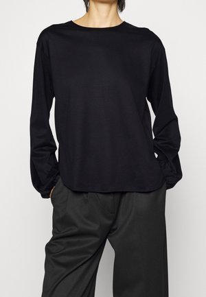 Osoba ubrana w czarną koszulę z długim rękawem z marszczonymi mankietami oraz luźne czarne spodnie, stojąca na jednolitym tle.