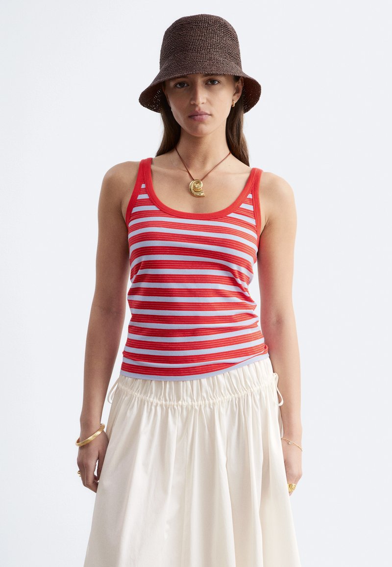 Jeune femme portant un chapeau seau marron, un débardeur rayé rouge et blanc, une jupe blanche et un collier en coquillage doré, regardant droit devant avec une expression neutre.