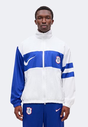 Homme portant un survêtement blanc et bleu Chelsea FC Nike avec la veste zippée, debout devant un fond clair uni.