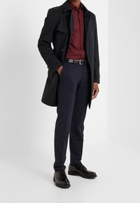 Zwarte trenchcoat over een donkerrode overhemd met knopen, gecombineerd met marineblauwe broek en zwarte schoenen. De jas heeft een dubbele knoopsluiting.