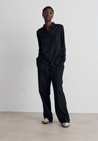 Black, pinstriped button-up shirt with two front pockets, paired with matching wide-leg trousers and silver slip-on shoes.