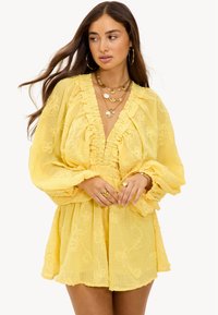 Femme portant une robe jaune brodée de fleurs avec des manches longues bouffantes et des colliers en or superposés, regardant de côté les mains jointes.