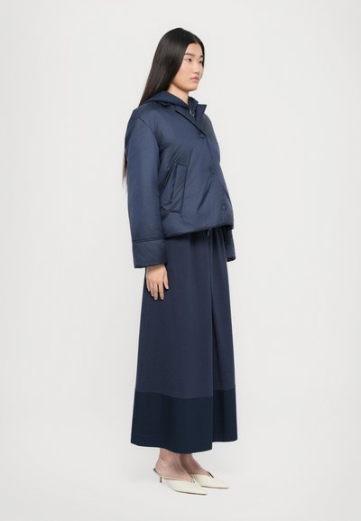 Temno modra skrajšana puhaste jakna s kapuco, ki ima dve sprednji žep in gumb zapiranje, nosi pa se jo čez dolgo temno modro obleko in bež čevlje.