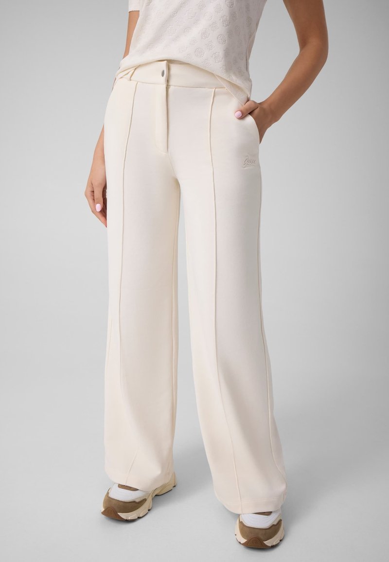 Femme portant un pantalon large couleur crème avec des coutures devant, associé à des baskets et un haut à manches courtes rentré.