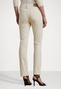 Lys beige, tettsittende jeans med baklommer og en lærlogo-patch på livet, sammen med svarte høyhælte sandaler.