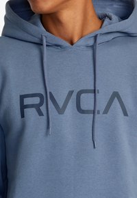 Felpa in misto cotone blu con tasca a marsupio frontale, coulisse e un logo scuro "RVCA" ben visibile sul petto. Tessuto morbido.