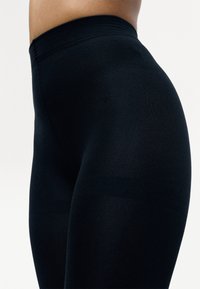 Nahaufnahme einer Person in hochtaillierten schwarzen Leggings, die einen glatten, eng anliegenden Stoff an Hüften und Oberschenkeln vor einem hellen Hintergrund zeigt.