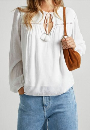 Vit texturerad blus med rund krage, dekorativ sömnad och knytning i detalj. Tyget har små upphöjda prickar. Kombineras med ljusblå jeans.