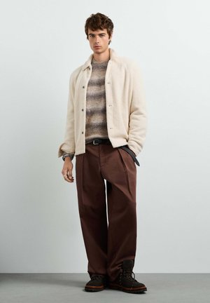 Young man wearing a cream buttoned jacket over a striped sweater, brown wide-leg pants, and dark boots, standing against a plain light wall.