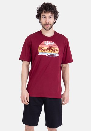 Homme aux cheveux bouclés portant un t-shirt bordeaux avec un motif coloré de coucher de soleil et de palmiers, et un short noir, debout devant un fond blanc.