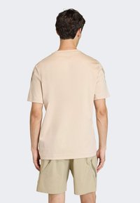 Camiseta de algodón beige con mangas cortas y cuello redondo clásico, mostrada en una persona que lleva pantalones cortos kaki claros. Sin gráficos ni patrones visibles.